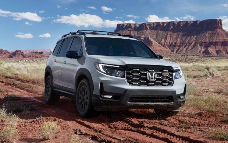 Front passenger-side view of the 2023 Honda Passport TrailSport in Sonic Gray Pearl with Honda Genuine Accessories, driving in the desert with a mesa in the background. 