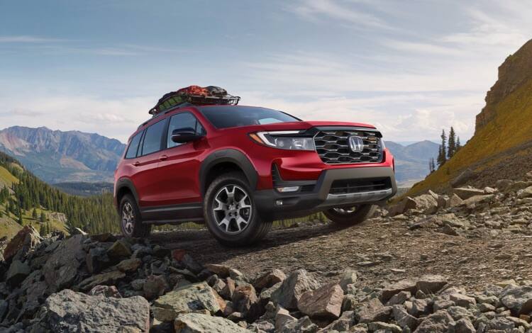 Passenger-side front view of the 2023 Honda Passport TrailSport in Radiant Red Metallic II, with accessory roof basket, driving on a rugged mountain road. 