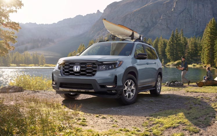 Driver-side front view of the 2023 Honda Passport TrailSport in Sonic Gray Pearl, with accessory kayak attachment, parked at a lakeside campground with male and female campers. 
