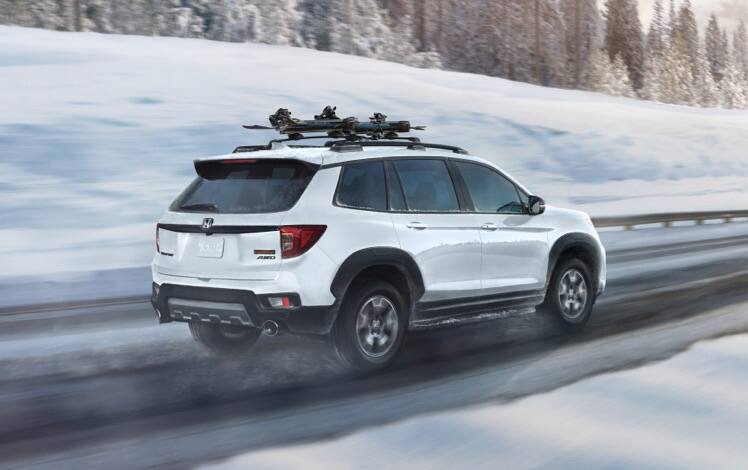 Passenger-side rear view of the 2023 Honda Passport TrailSport in Platinum White Pearl with accessory ski/snowboard attachment, driving on a snowy mountain road. 